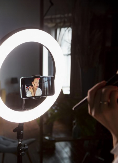 Fotografia d`un cèrcol de llum i un telèfon mòbil, a la pantalla del qual es veu una jove gravant-se mentre es maquilla.
